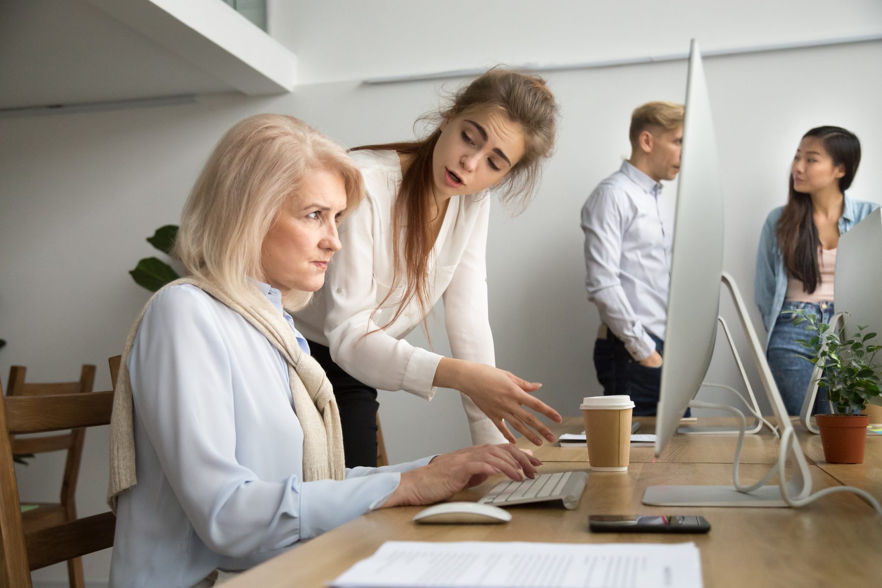 An older woman sitting at a desk using a computer, with a younger colleague leaning over and speaking to her in an office setting, symbolising workplace age discrimination.