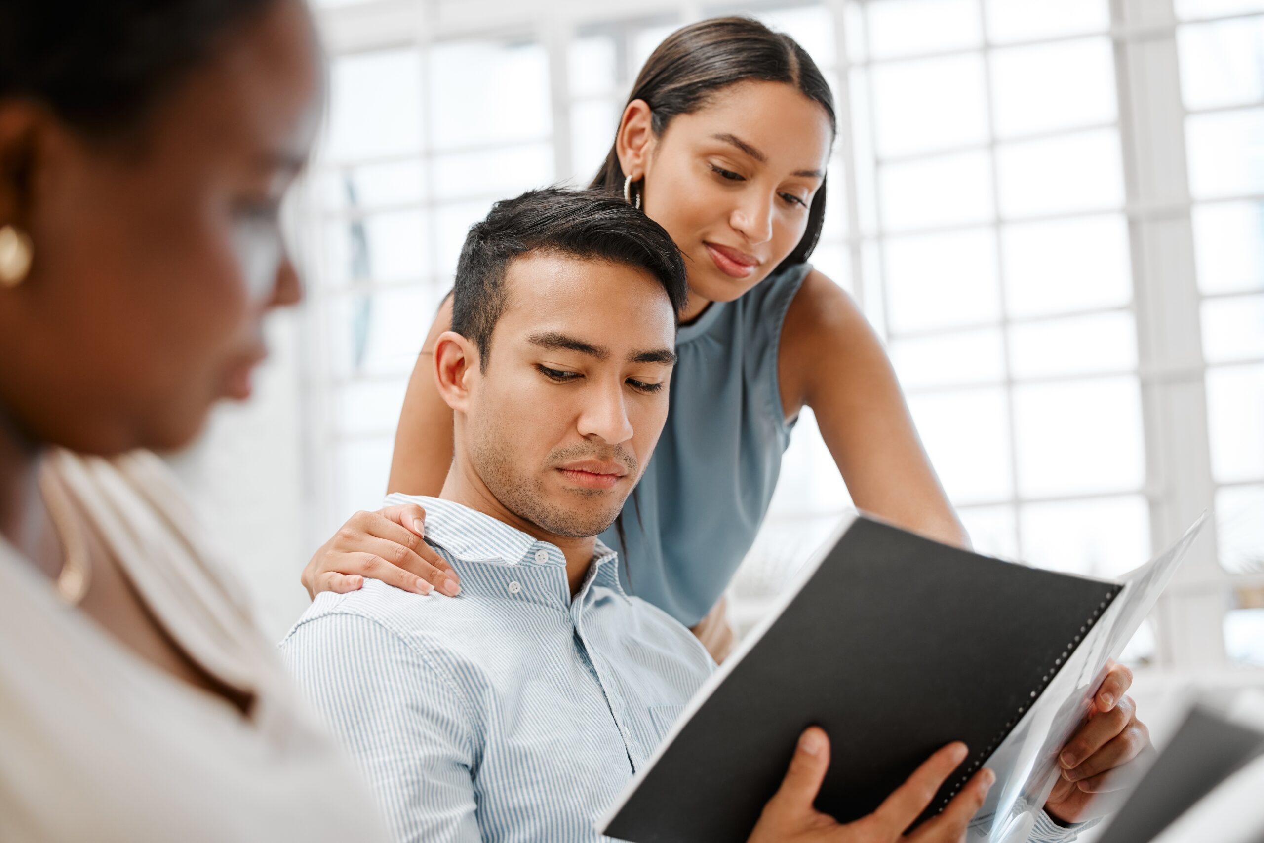 A stressed businesswoman sits at a desk with her head in her hand, symbolising workplace harassment, while two colleagues stand in the background.